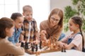 Happy female teacher in glasses pointing at board and explaining moves to cheerful children during chess lesson in daytime at school